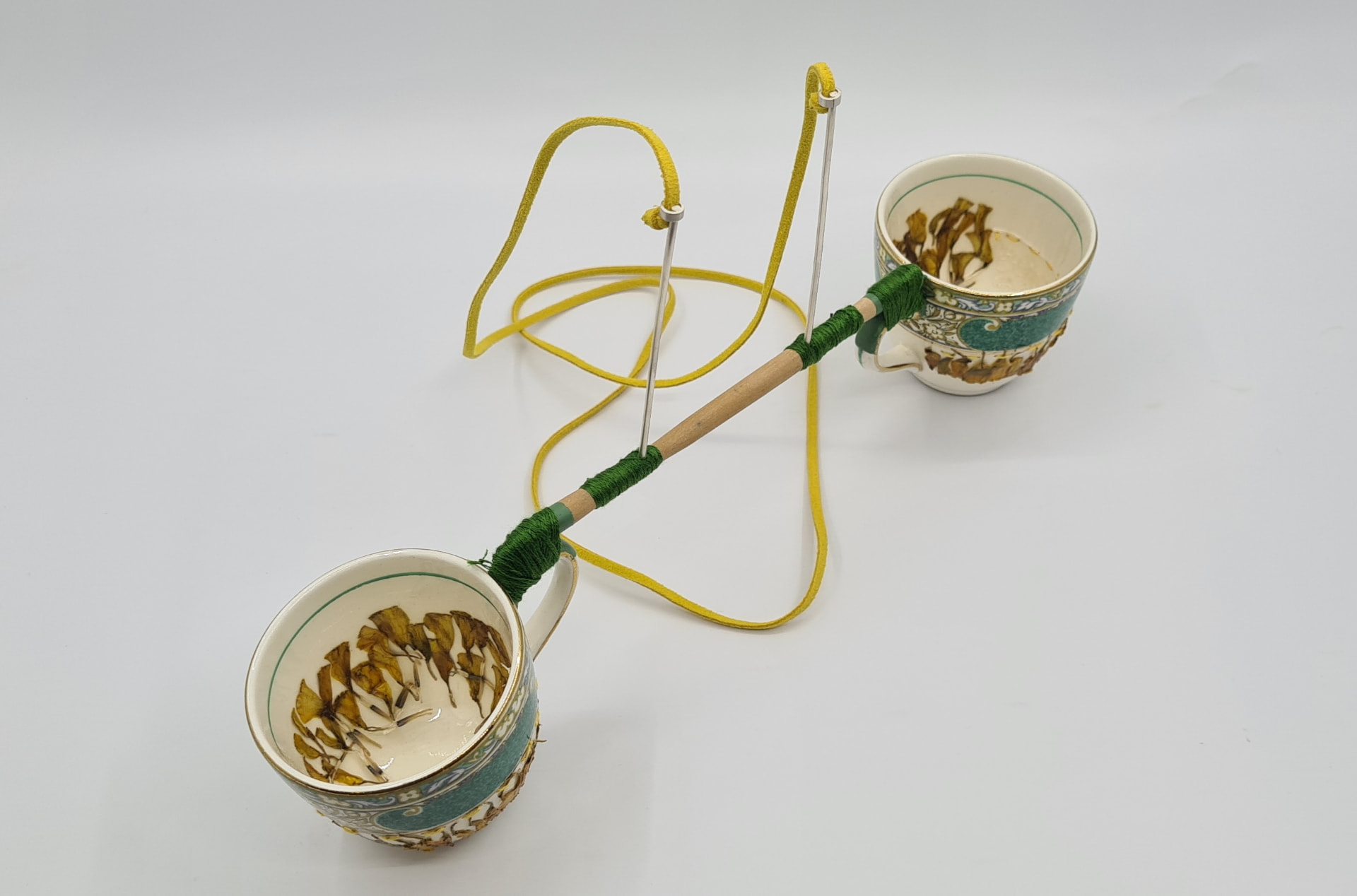 Neckpiece made from two heirloom porcelain teacups holding pressed leaves, joined by a thread-wrapped wooden rod on a yellow suede cord.