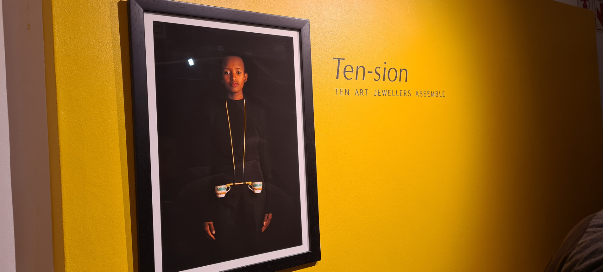 TEN-SION exhibition wall at Oude Leeskamer Gallery — framed portrait of a woman wearing a porcelain cup necklace beside the show title on a yellow wall.