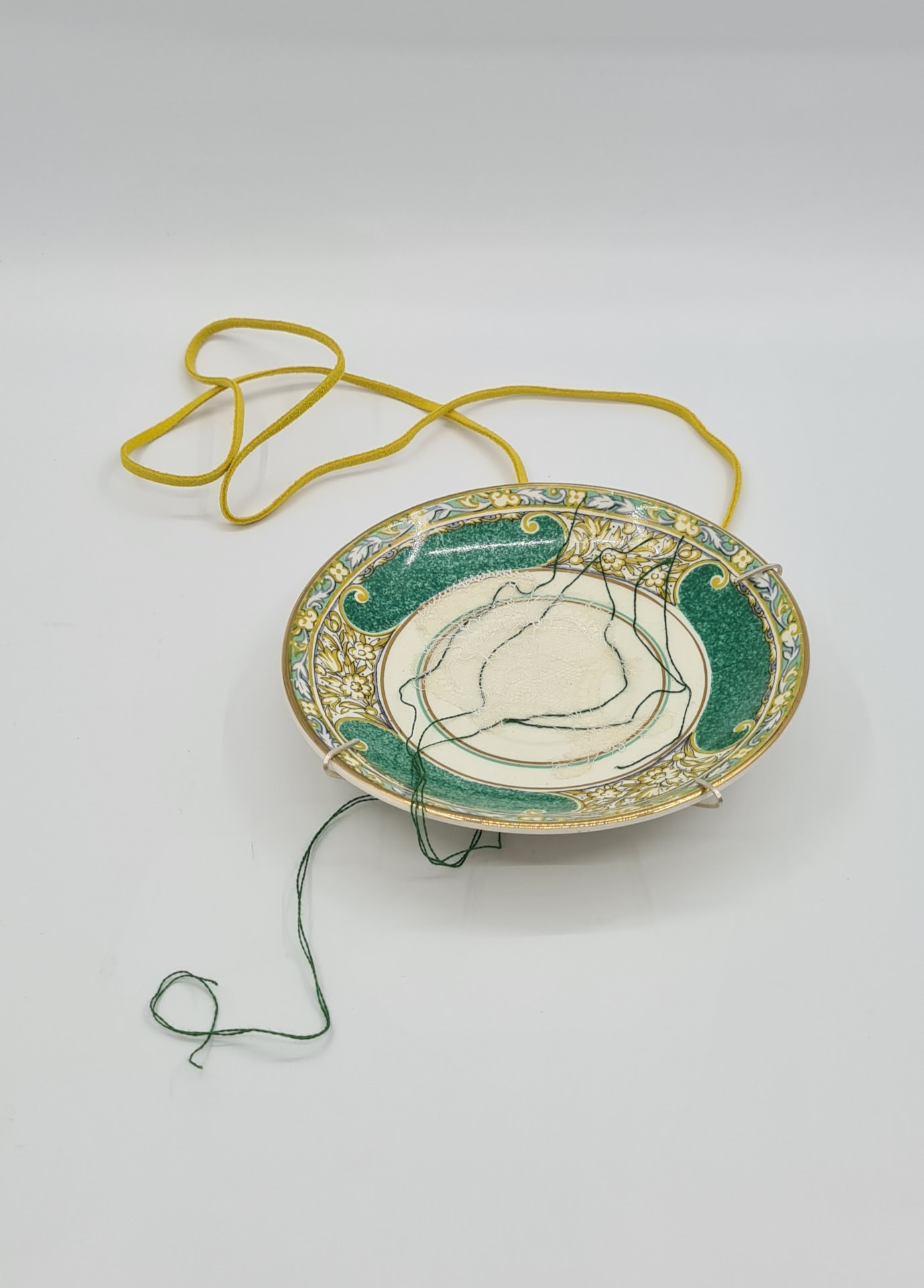 Neckpiece made from a green and gold patterned porcelain saucer stitched with dark green thread on a yellow suede cord.